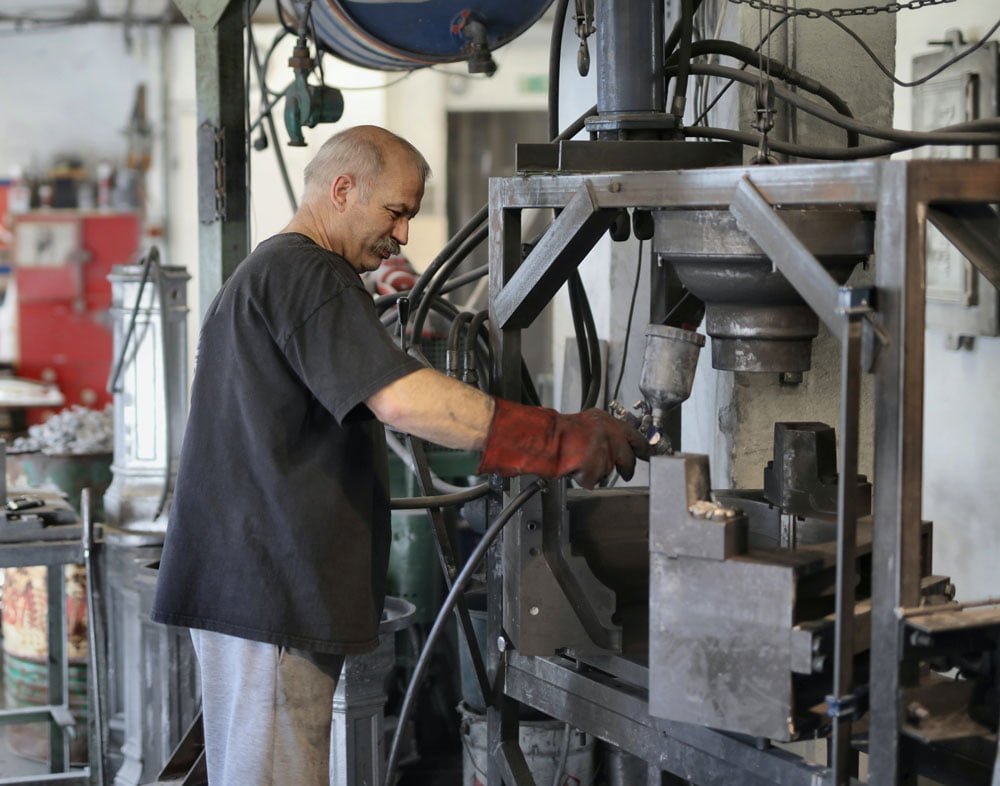 Man in a workshop operates industrial machinery while wearing gloves and a dark t-shirt.