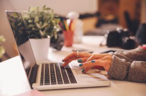 Hands typing on a laptop keyboard at a desk with a potted plant and colorful pens in the background.