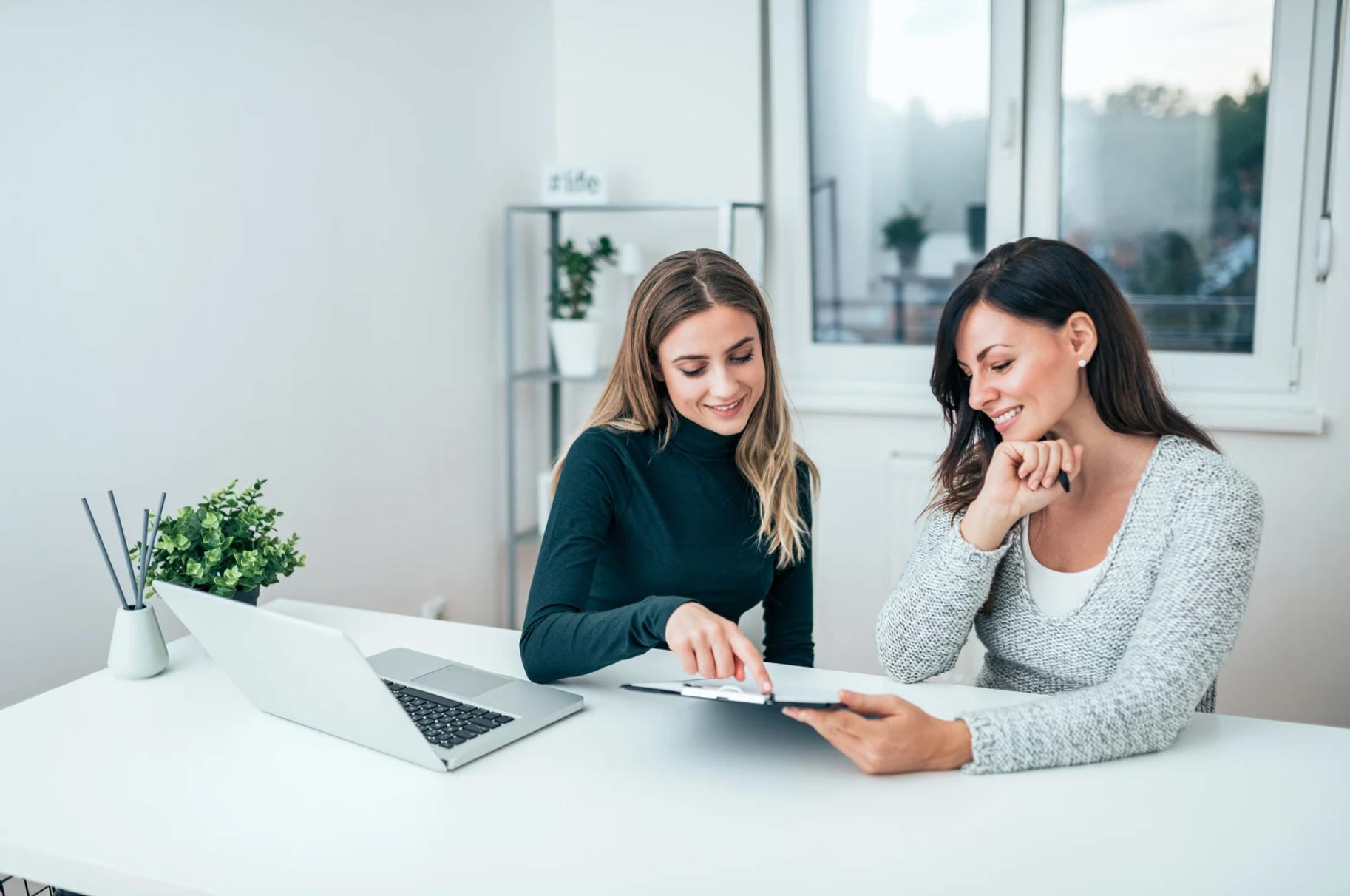 Two women sitting at a desk, smiling and looking at a clipboard, with a laptop nearby.