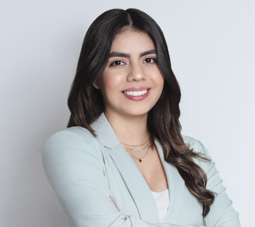 Woman with long dark hair in a light blue blazer, smiling with arms crossed against a light background.